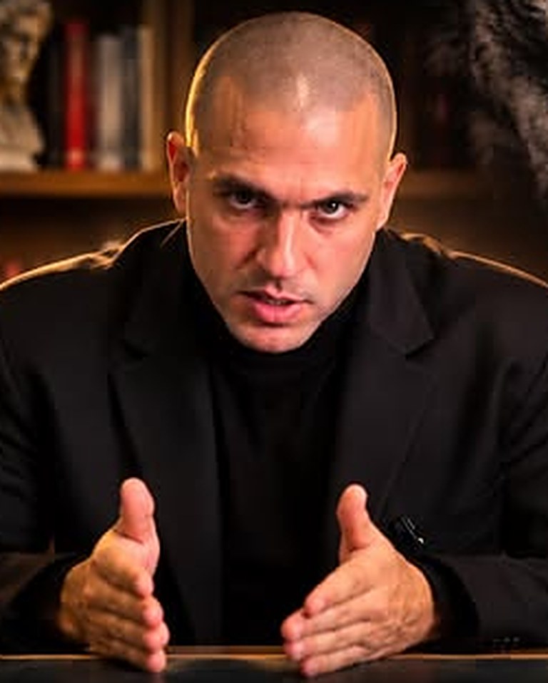 Jay, the Maskless Wolf, in a black suit at his desk in a book-lined study